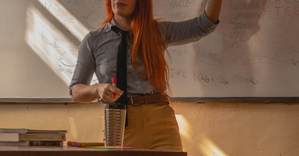 A teacher with long red hair gives a lesson while pointing at a whiteboard in a sunlit classroom.